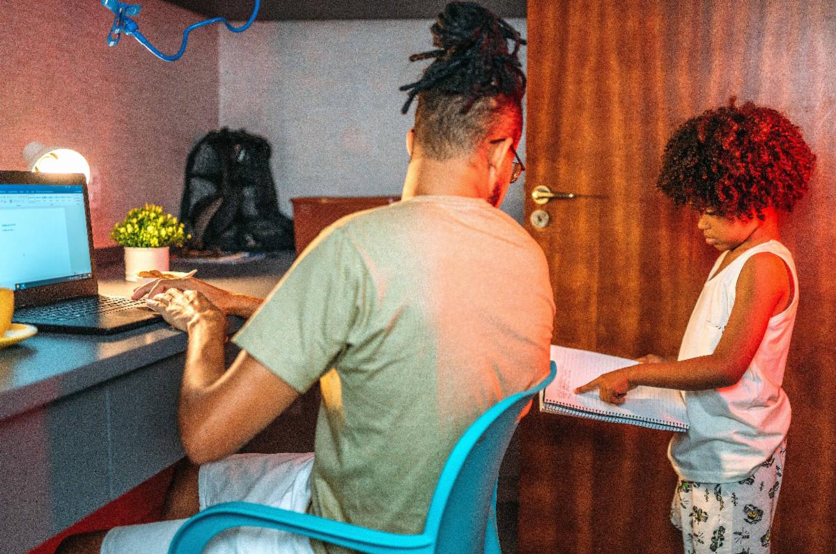 A young child points to a list on a piece of paper while a parent turns from thier laptop to look