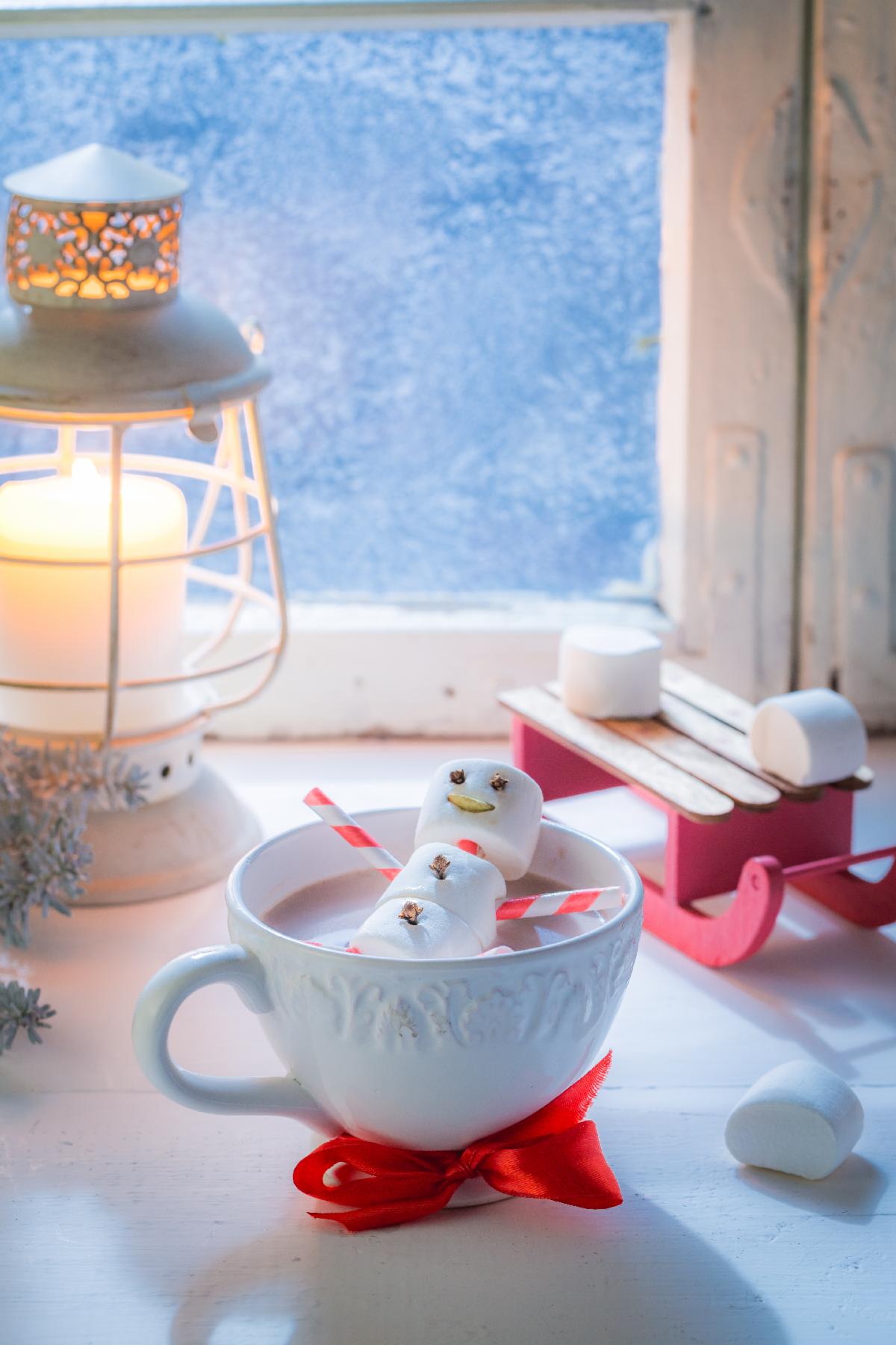 A marshmallow snowman floats in a cup of hot chocolate while a candle burns near a frosted window