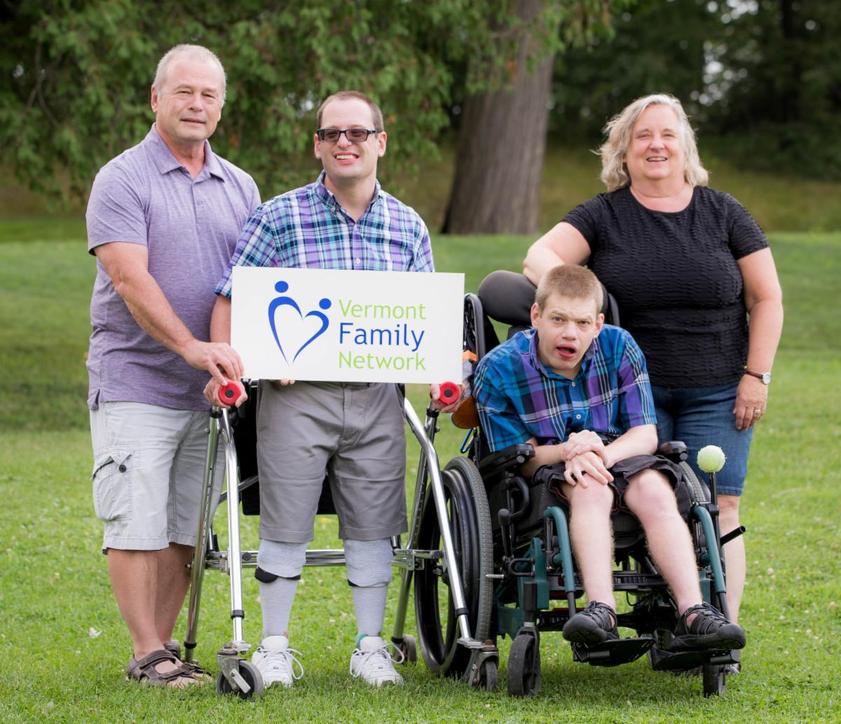 Two parents stand beside their two adult children who hold a VFN sign, one who uses a wheelchair and the other a stander