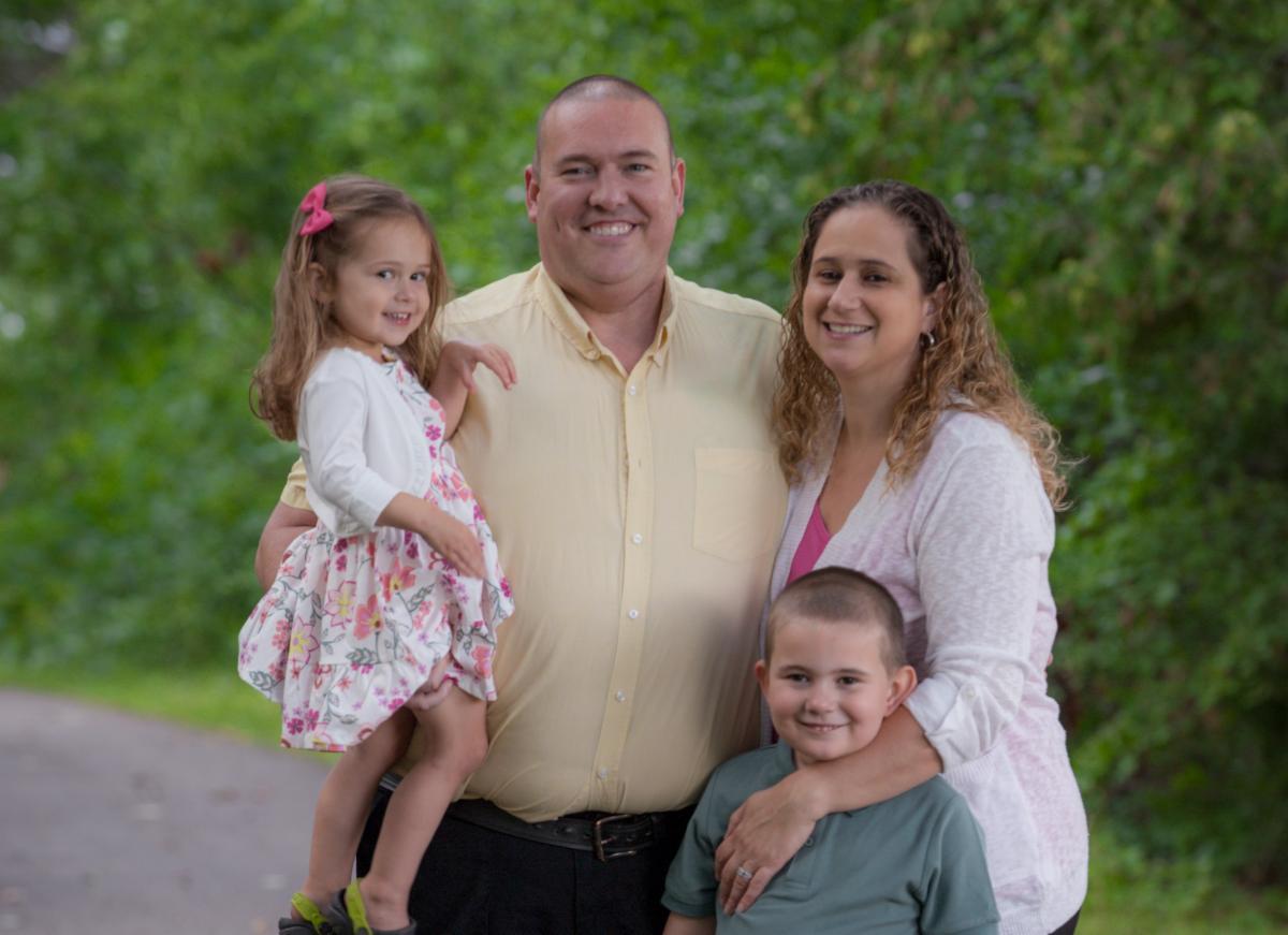 A family with two parents and two children wearing white, yellow and green, stand in front of a green wooded background