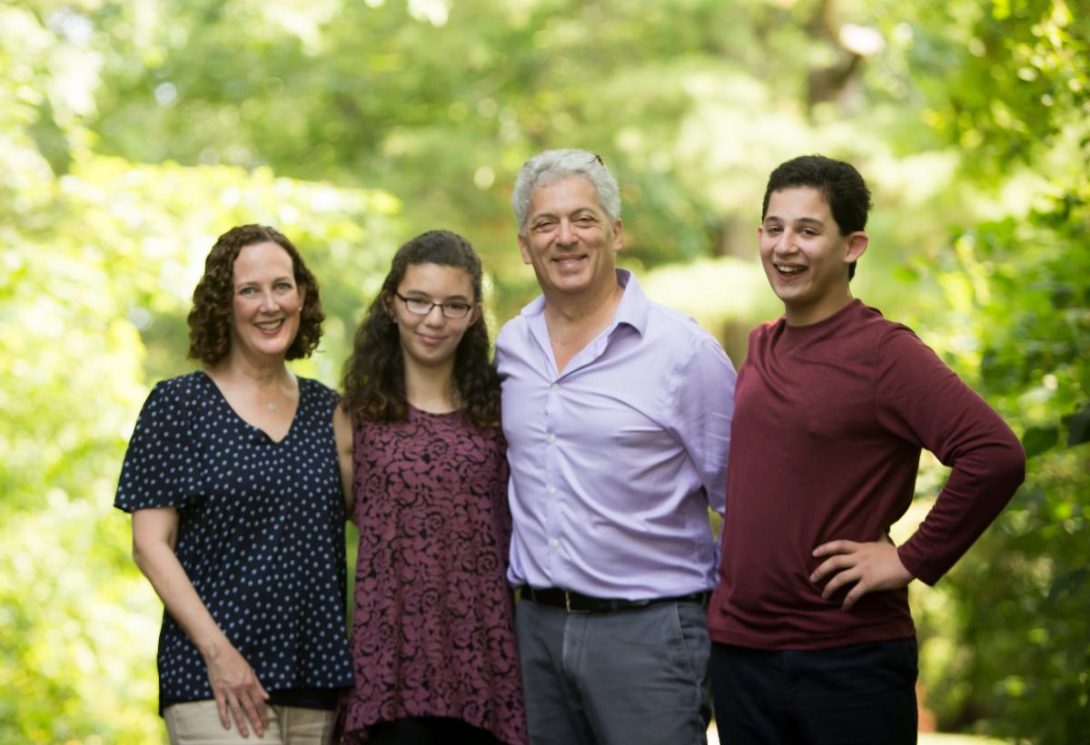 A family with two young adult children stand in front of a background of cascading green tree leaves