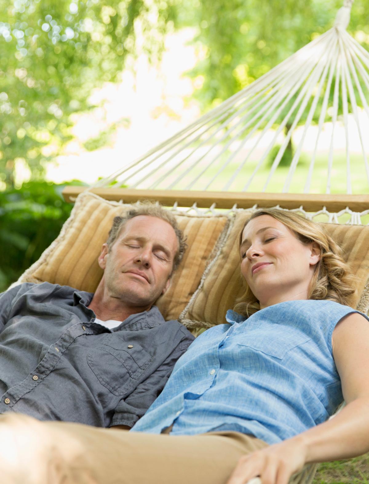 A couple with eyes closed rest together in a hammock in the shade of green trees