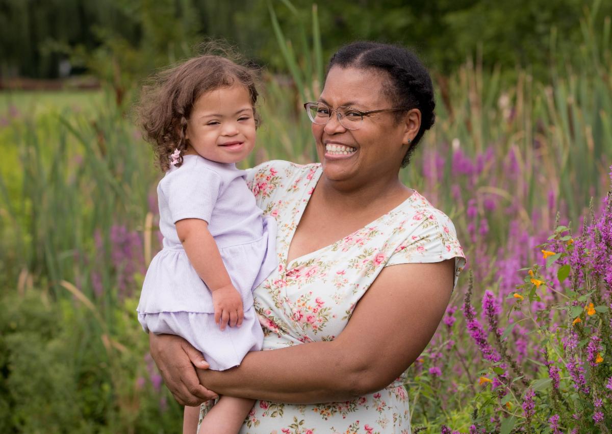 A parent hold toddler both wearing white dresses smile in front of a meadow of purple flowers