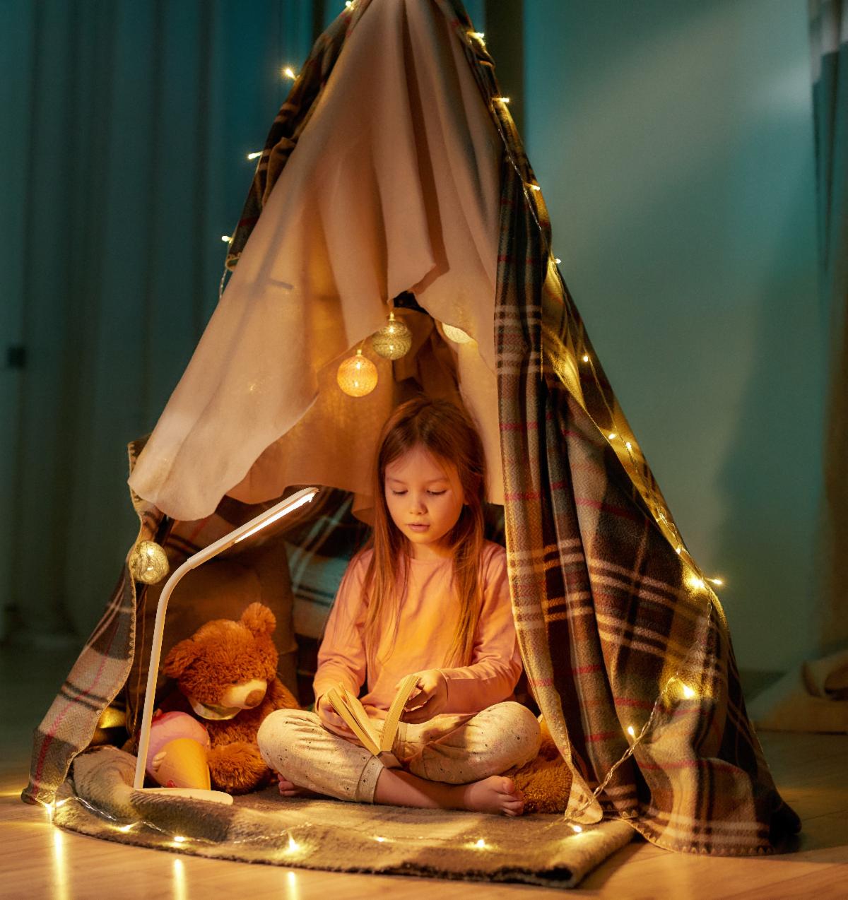 Child sits and reds a book inside of a tent made of blankets and decorated with fairy lights