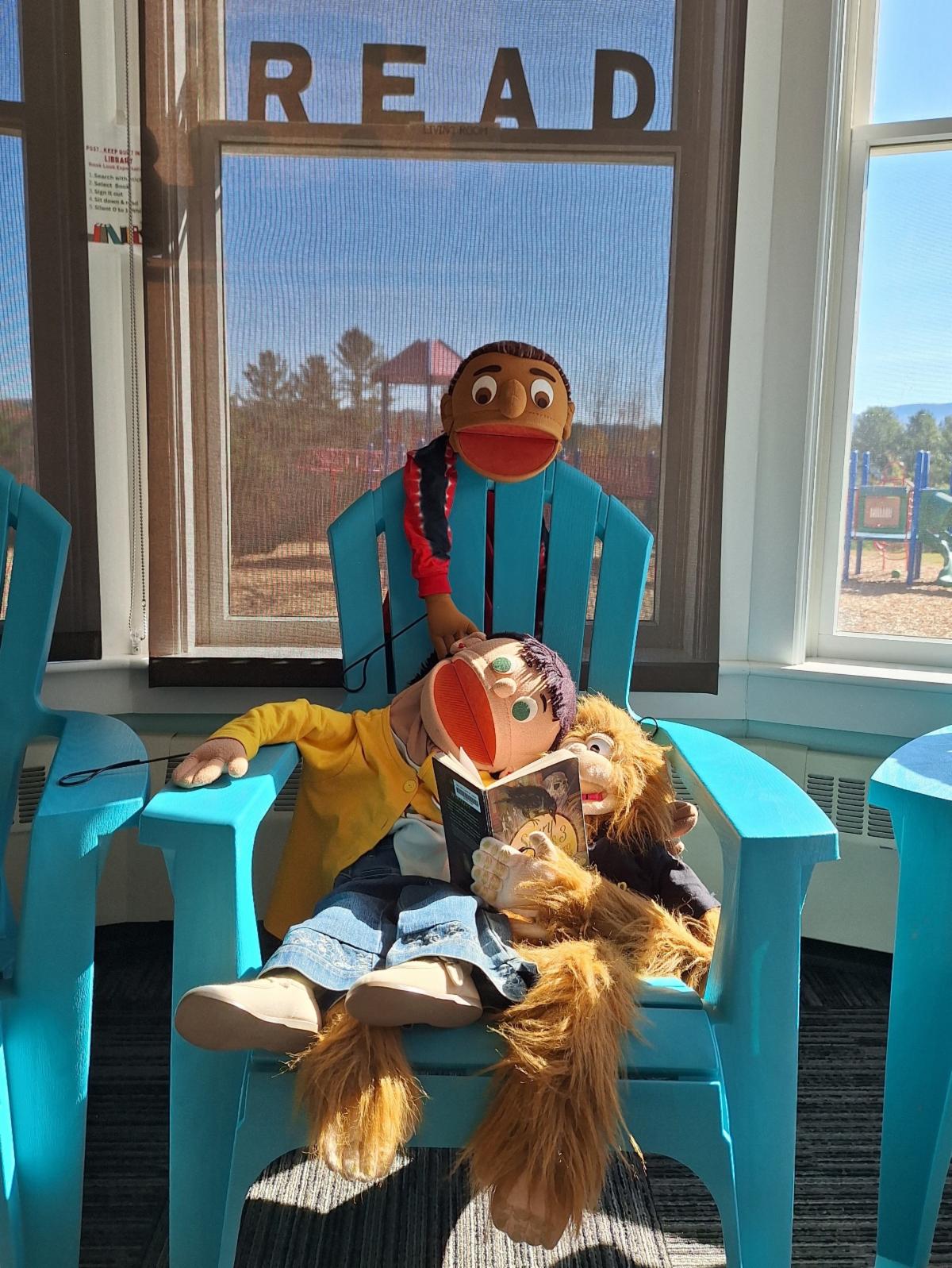 A group of puppets sitting in a blue Adirondack chair read a book with the word "read" on the wall behind them.