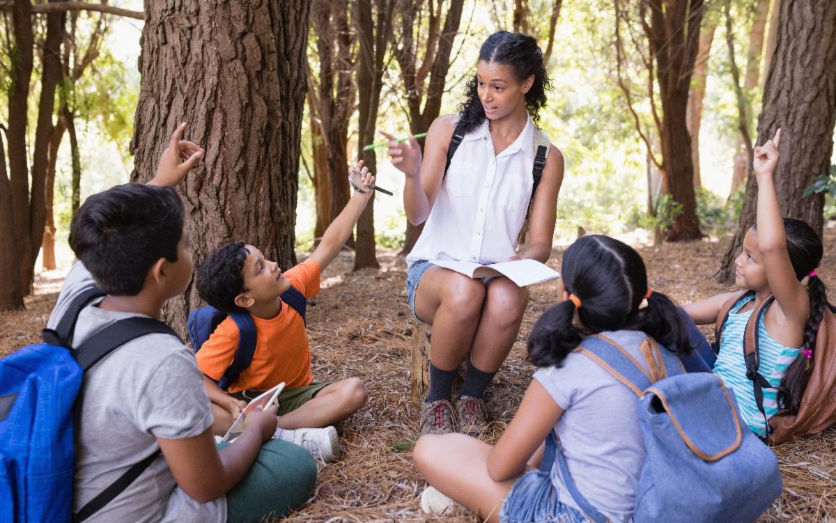 A group of children sitting on the ground with their summer camp leader in the woods