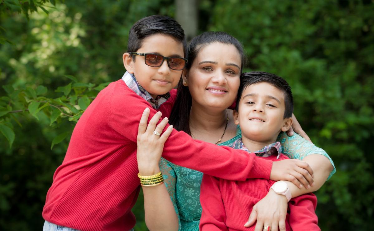 A parent wearing a green dress is hugged by their two children who are wearing red sweaters, one child is wearing dark glasses.