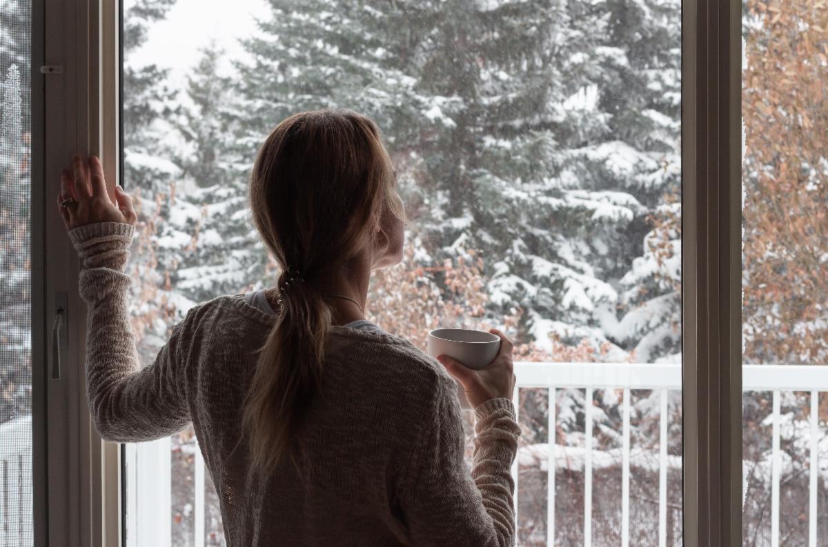 A parent holding a cup of tea stands in front of a large glass door looking out at a winter scene with snow on the trees beyond