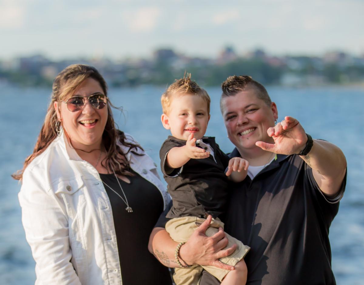 Two smiling parents hold their toddler while one parent and their child lift thier hands forward in a waving gesture