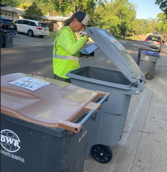 organics cart at curb with brown lid, man in yellow vest opening trash cart with grey lid