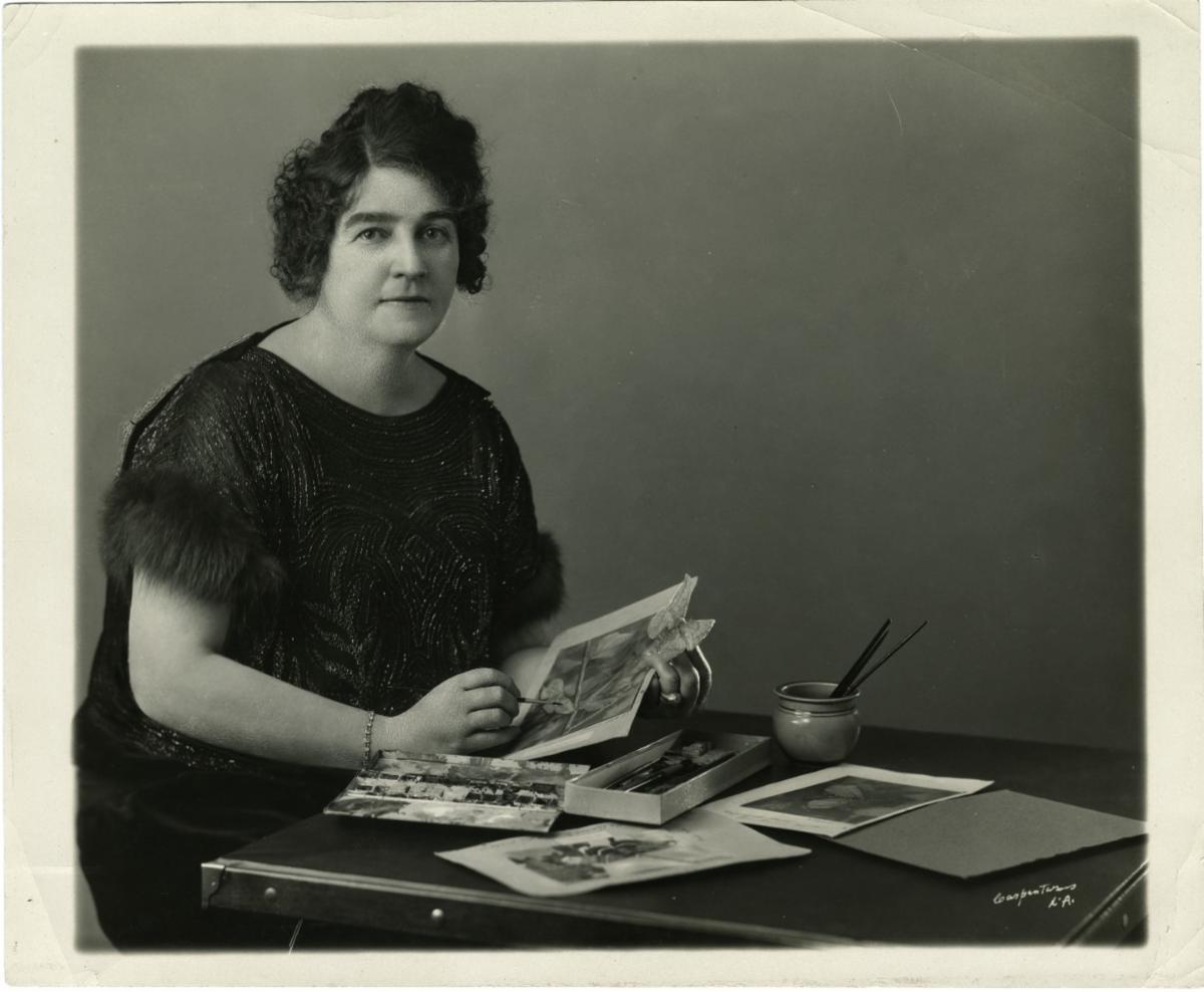 Black and white portrait of American author and naturalist Gene Stratton-Porter seated at a table, holding a small artwork and surrounded by paintbrushes, a palette, and prints.