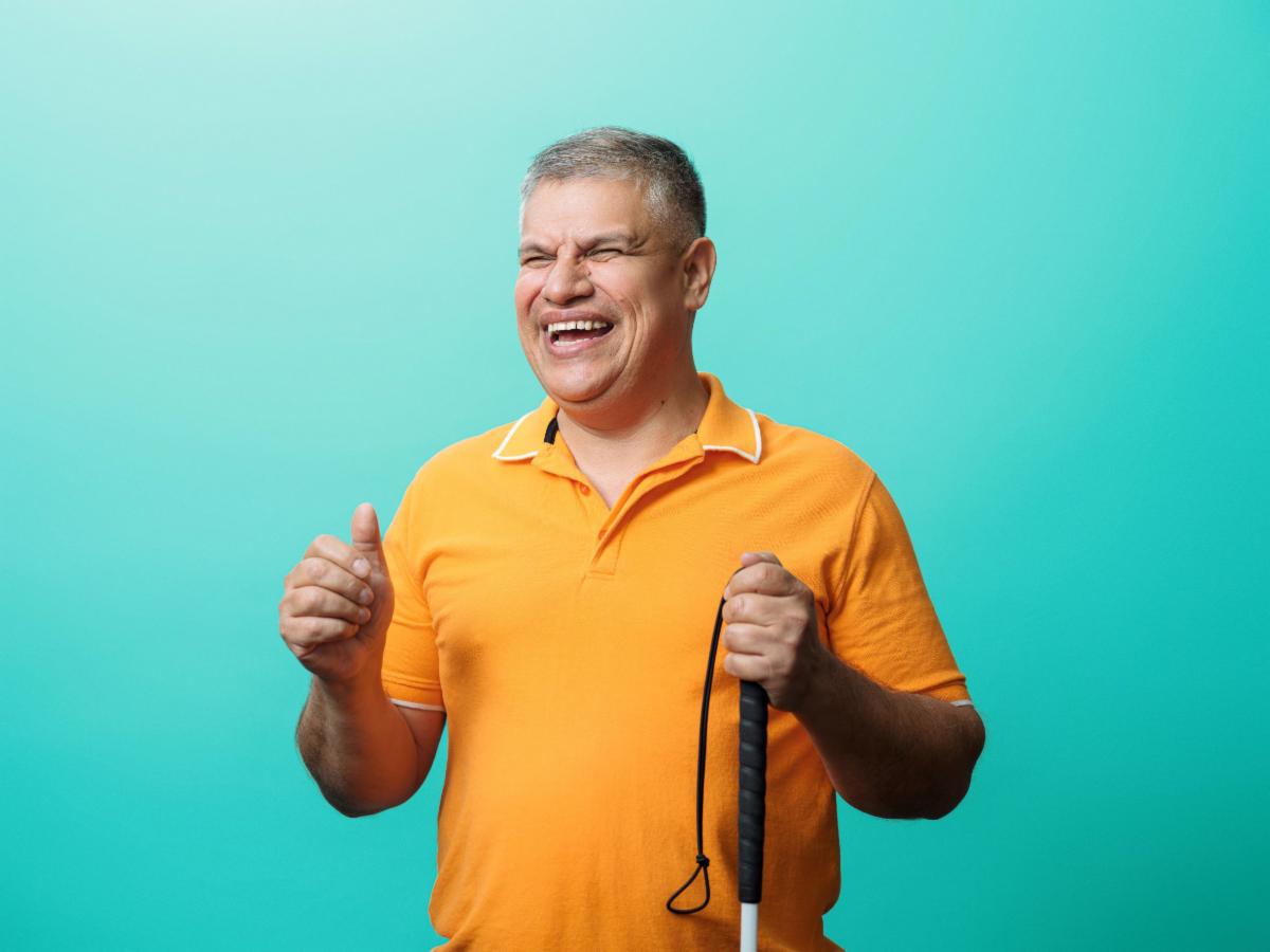 Smiling older man in an orange polo shirt holding a white cane against a bright turquoise background.