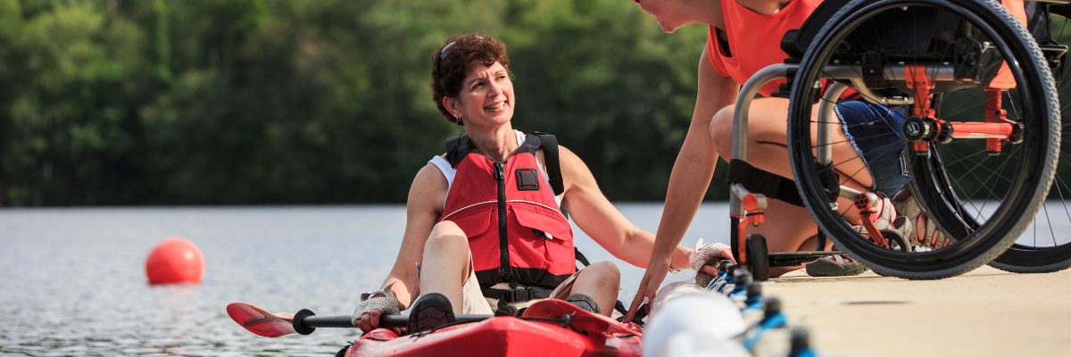 A woman with a spinal cord injury is in a kayak on a lake. Her wheelchair is on the dock. She is speaking with a kayak instructor. 