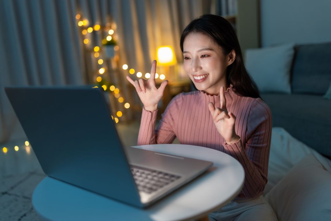 Woman is using ASL to communicate over video chat on a laptop at home. Christmas lights are in the background.