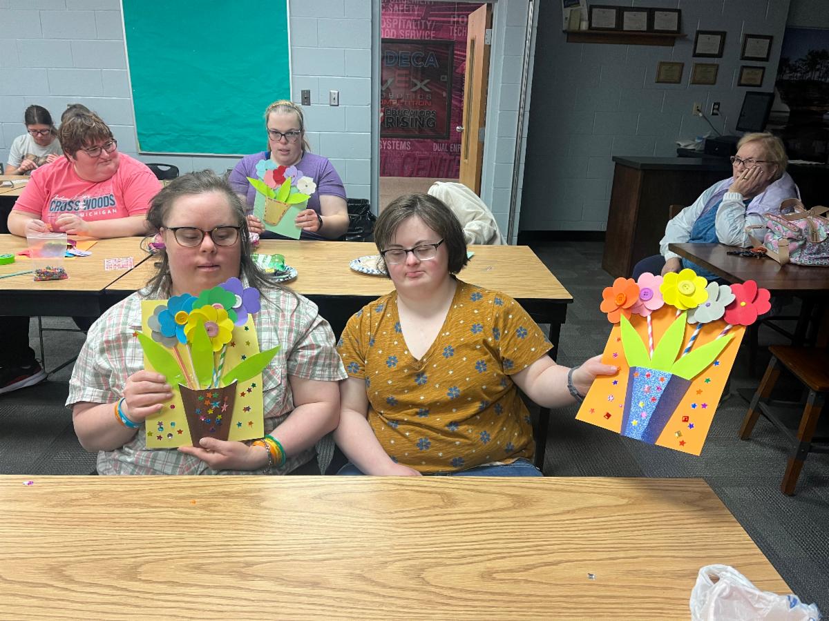 Two women with intellectual disabilities display the floral art projects they created in a group art class. Other students are seated at tables behind them working on similar art projects.