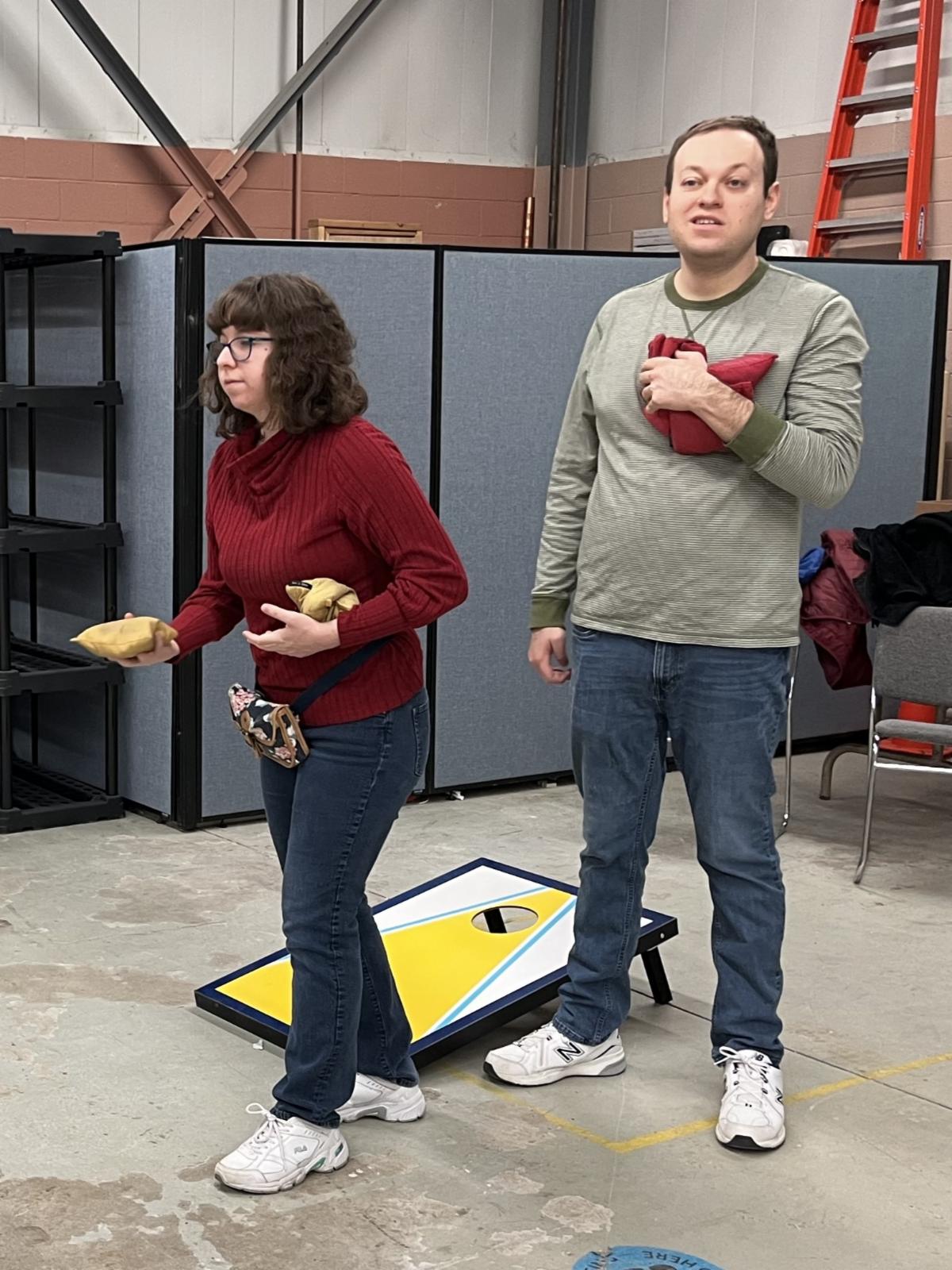Two people holding bean bags are playing cornhole at an indoor recreation event.