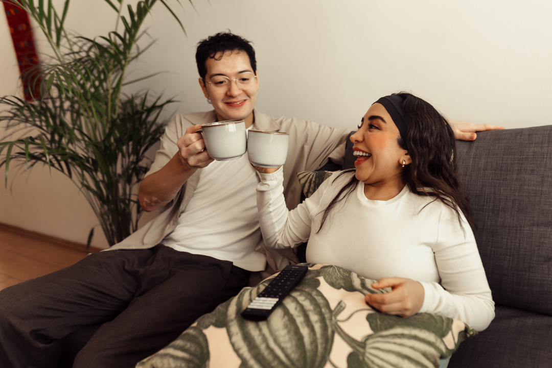 Two people are sitting on a sofa clinking coffee mugs in celebration.