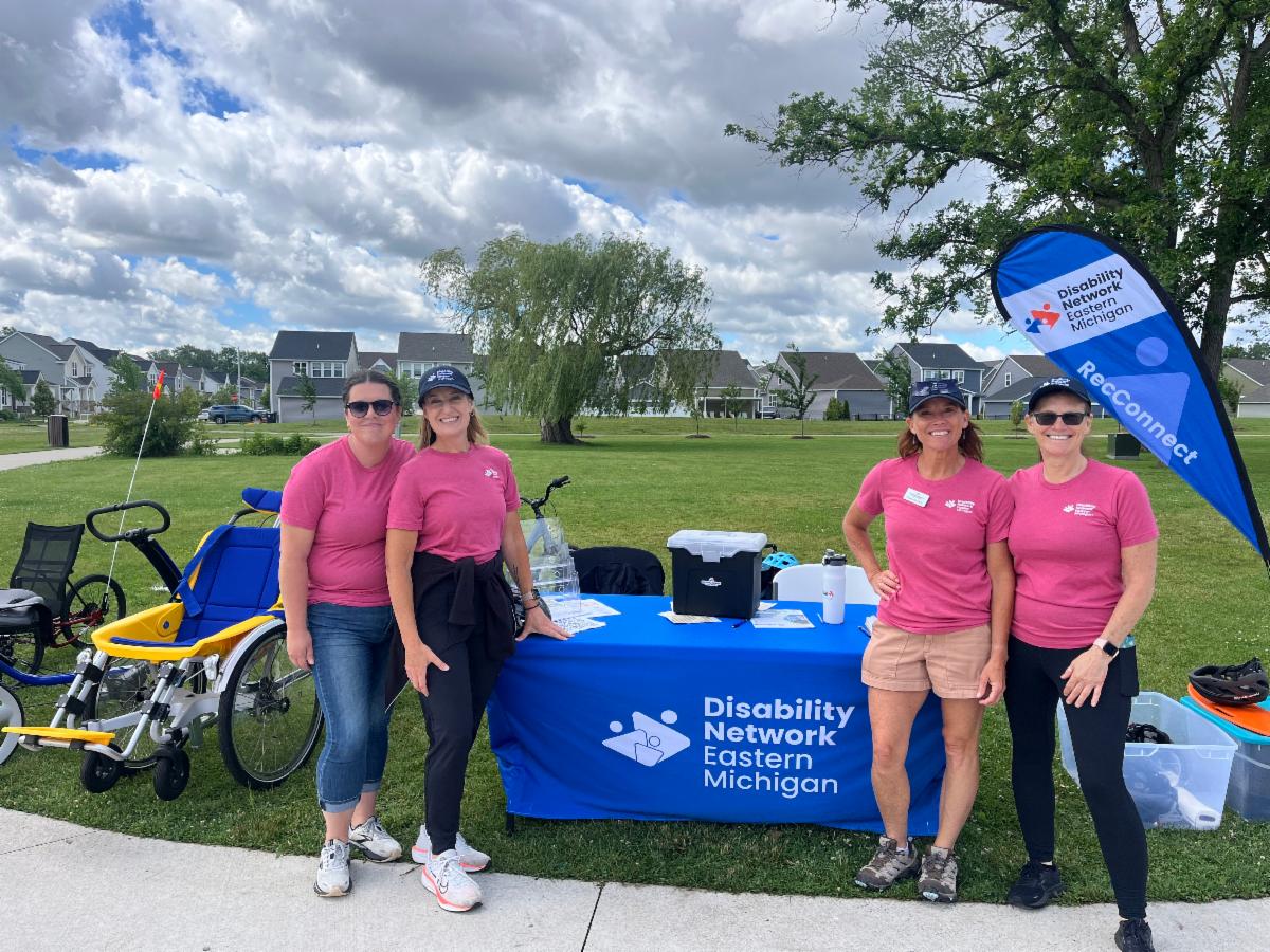 Smiling Disability Network Eastern Michigan staff stand in front of a registration table next to adaptive bikes, ready to greet adaptive recreation participants.