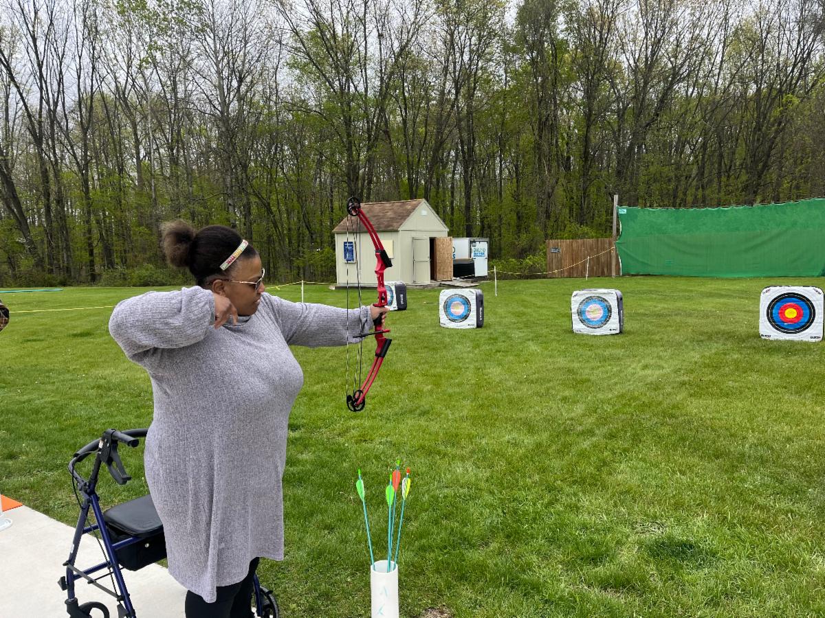 A woman is getting an adaptive archery lesson at at outdoor range. She is standing next to her rollator.