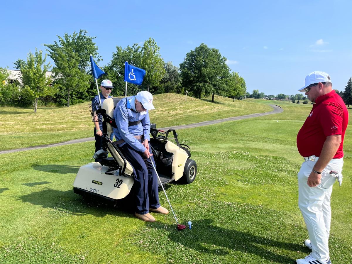 A man using an adaptive golf cart prepares to tee off while two other people watch.