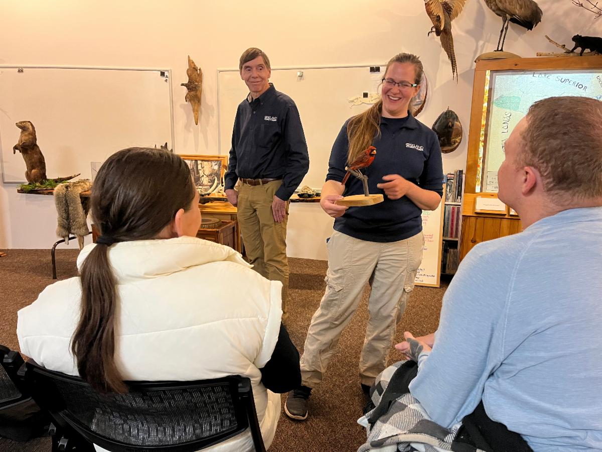 A park naturalist stands indoors holding a mounted bird taxidermy while speaking to a small seated group. Animal exhibits and educational displays line the walls behind them.