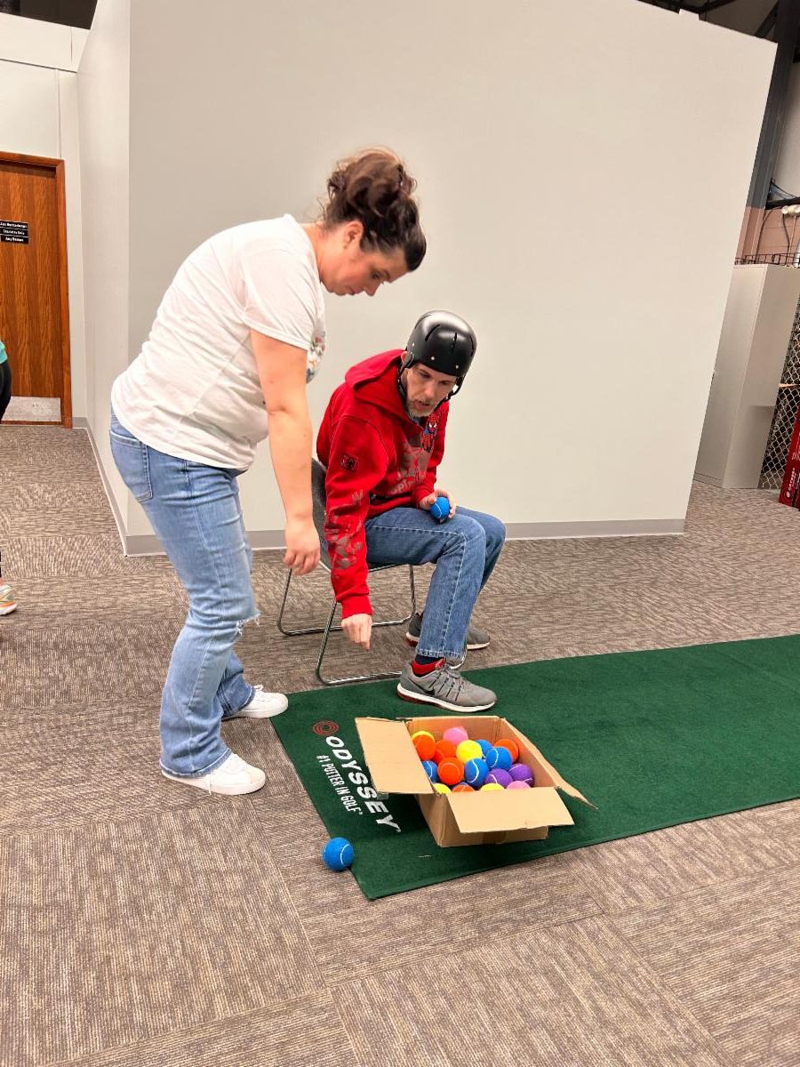 Seated man is reaching for tennis balls to throw at at velcro target. Staff is assisting.