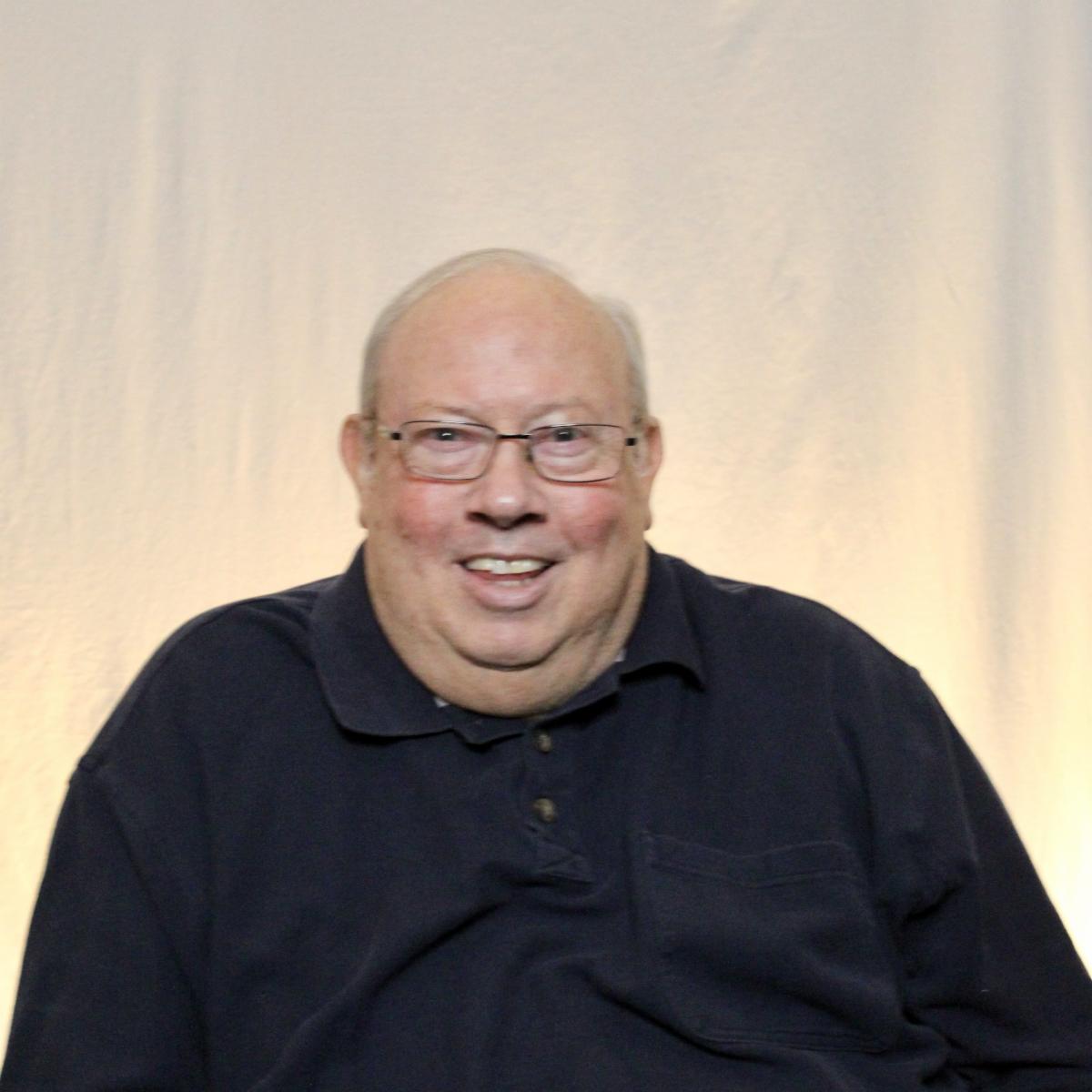 Randy Charon. A balding white man with a big smile. He is wearing a black polo shirt and glasses.