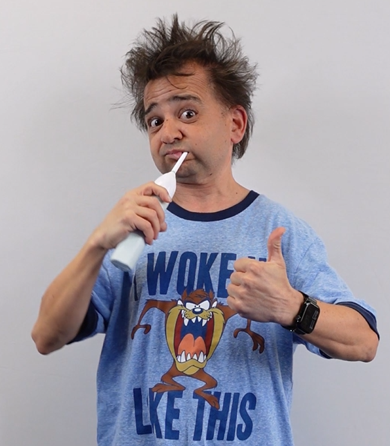 Screenshot from a Disability Network Eastern Michigan YouTube short. Staff Jeffrey Astrein gives a thumbs up as he brushes his teeth with an electric toothbrush. He has bedhead and is dressed in pajamas that say I woke up like this.
