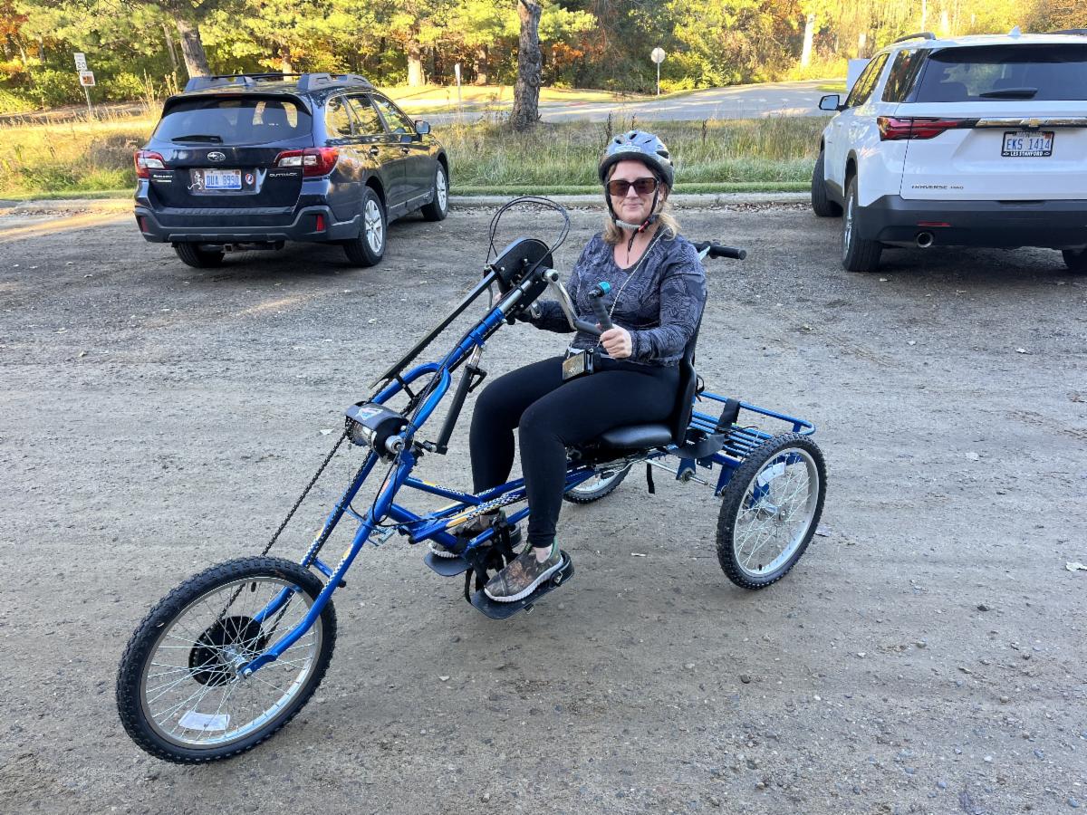 Woman wearing a bike helmet prepares to ride a handcycle recumbent trike in gravel park parking lot.