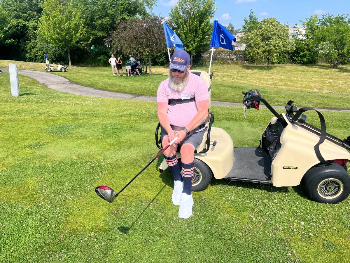 A man is using an adaptive golf cart to help him stand when hitting a golf ball.