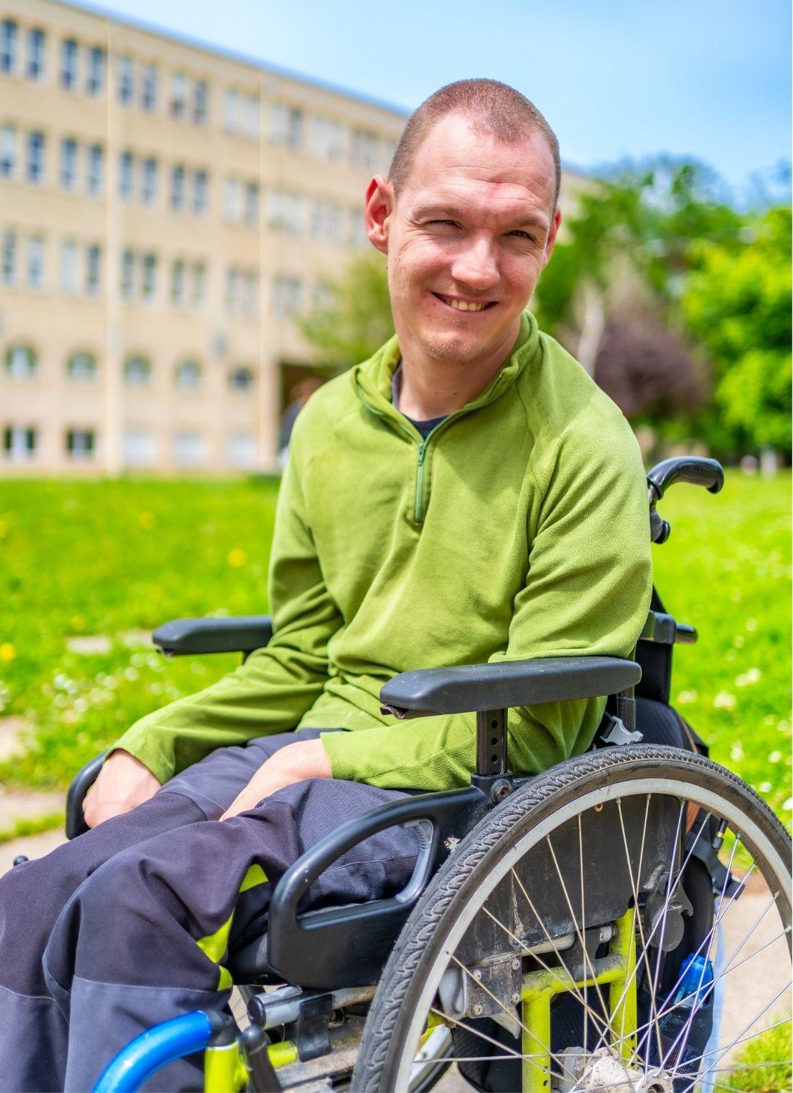 A smiling man with cerebral palsy is seated in a wheelchair outside a building on a college campus.