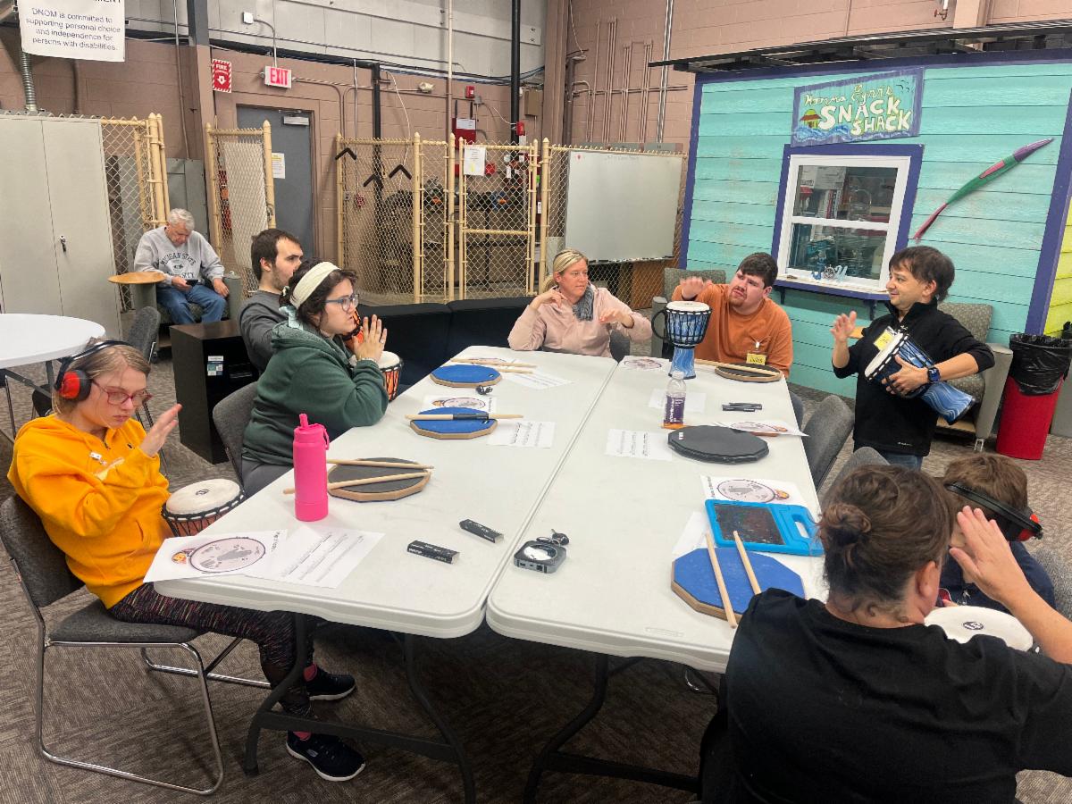 An instructor stands at the front of a group, holding a hand drum while participants seated around the table raise their hands in a drumming activity. A couple of attendees wear headphones or earmuffs to reduce sensory overload.