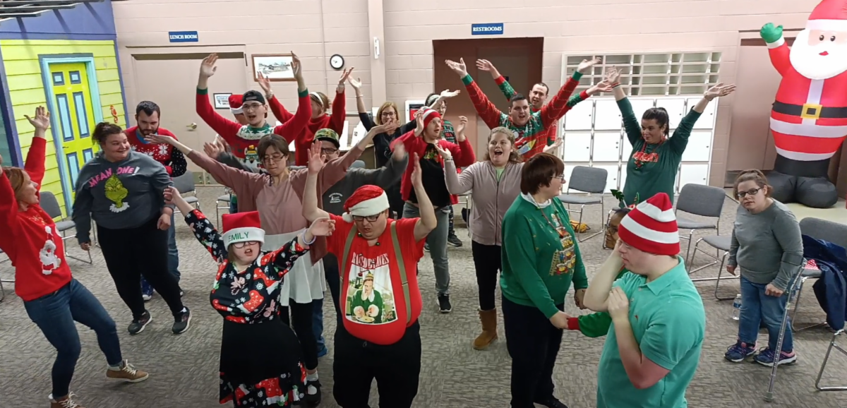 A diverse group of people dressed in holiday outfits are dancing together at Disability Network Eastern Michigan's Holiday Karaoke Party.