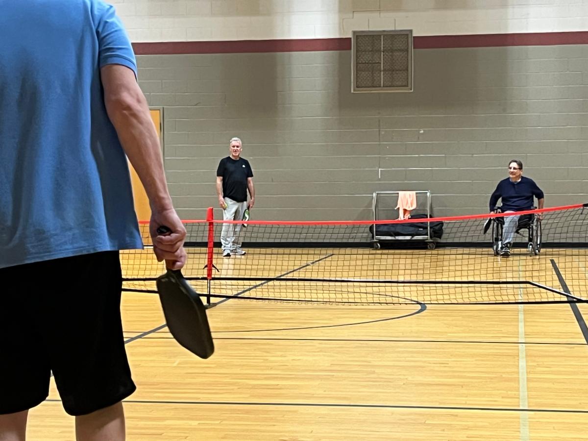 Three players are preparing for a game of pickleball in a gym.