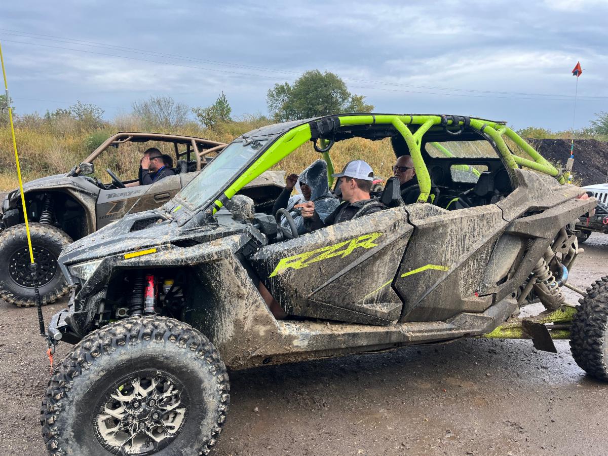 People prepare to drive and ride in two muddy off road vehicles.