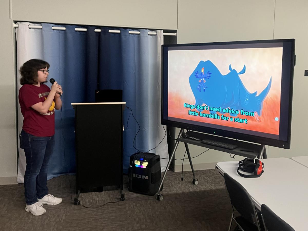 A woman is singing karaoke into a microphone while reading the lyrics on a large screen.