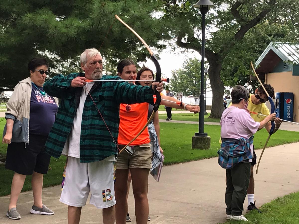 Two people are learning adaptive archery while bystanders watch.