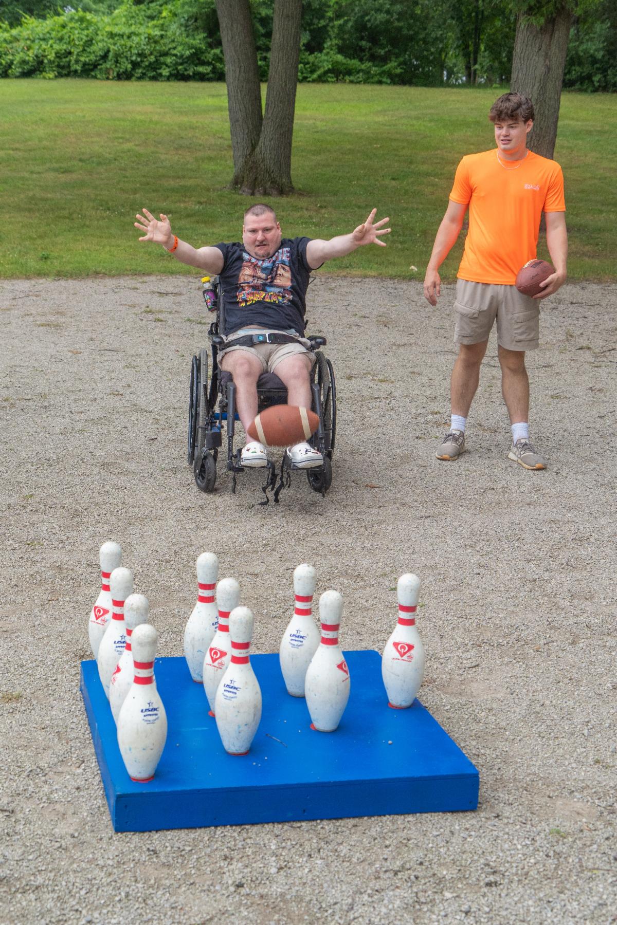 A man using a wheelchair throws a football at bowling pins in a park as another man holding a football watches.
