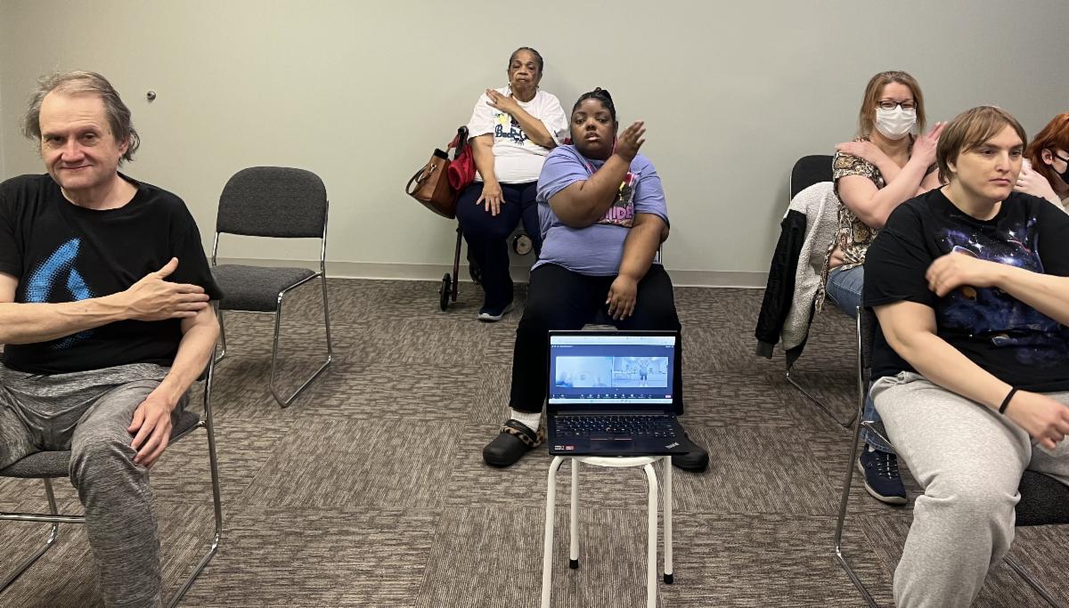 People seated in rows are participating in a chair exercise class. A laptop if focused on the instructor, broadcasting live to participants at home.