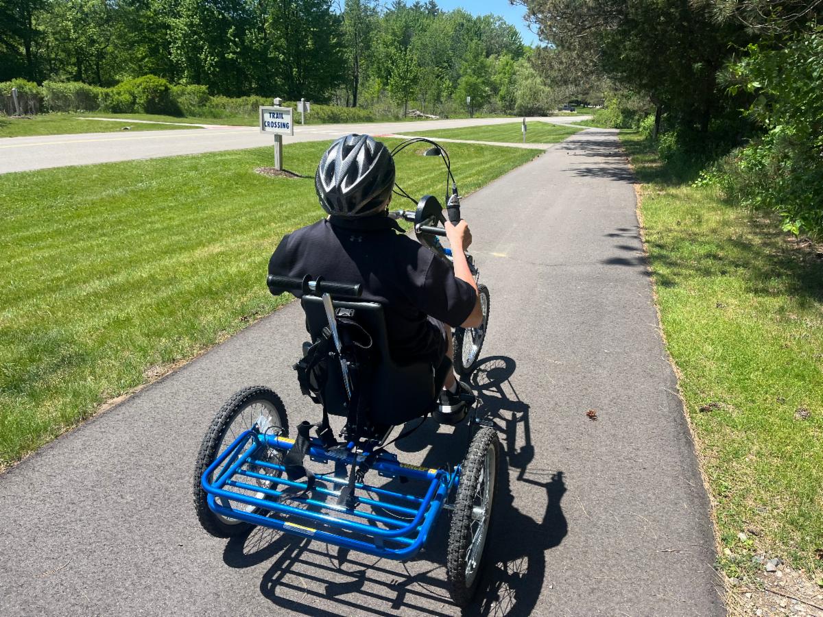 A person wearing a bike helmet is riding a handcycle trike on a paved park path.