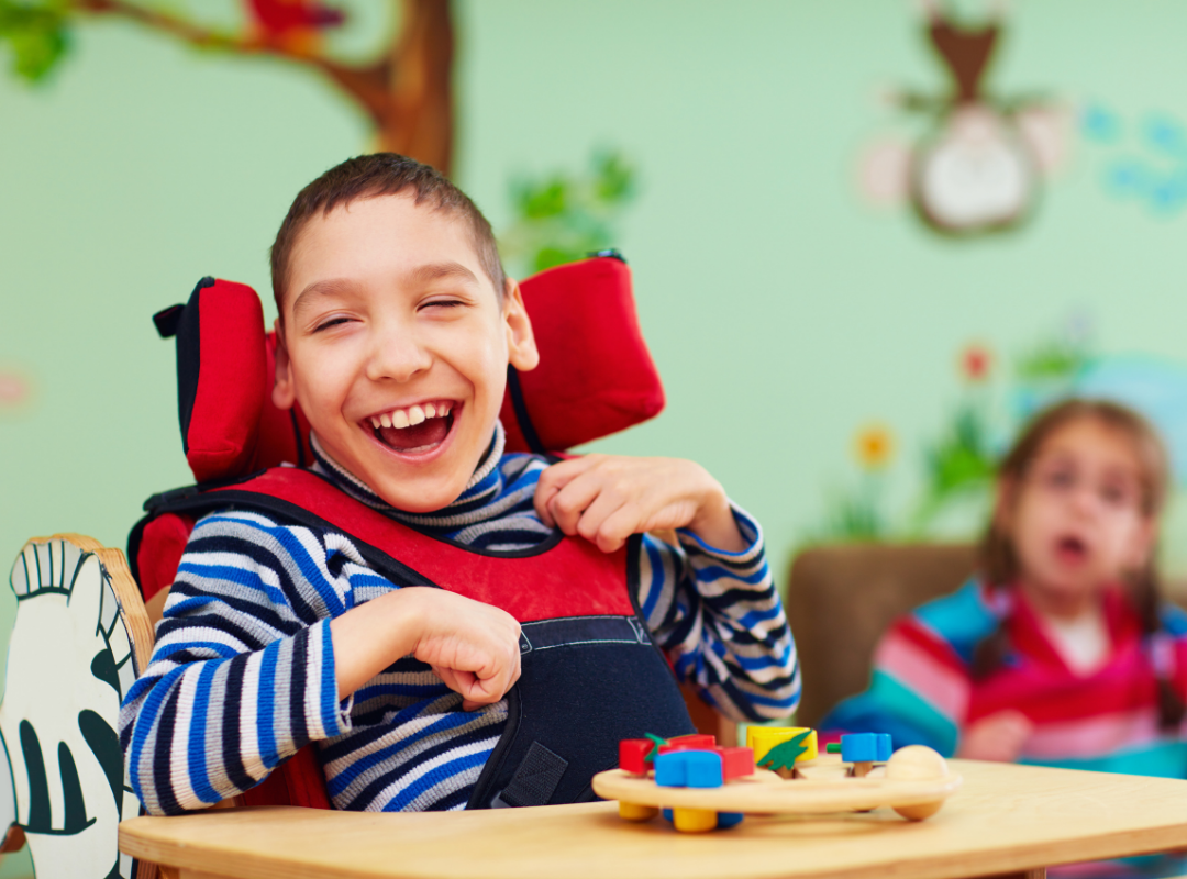 Laughing boy using a wheelchair.  His sister is in the background.