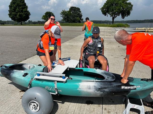 A wheelchair user wearing a lifejacket approaches a kayak on a kayak cart at a boat launch. Disability Network Eastern Michigan staff help stabilize the kayaking and a transfer bench. 