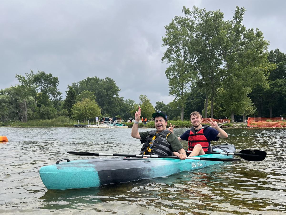 Two people wearing life jackets in a tandem kayak pause from paddling to smile and wave for a photo.