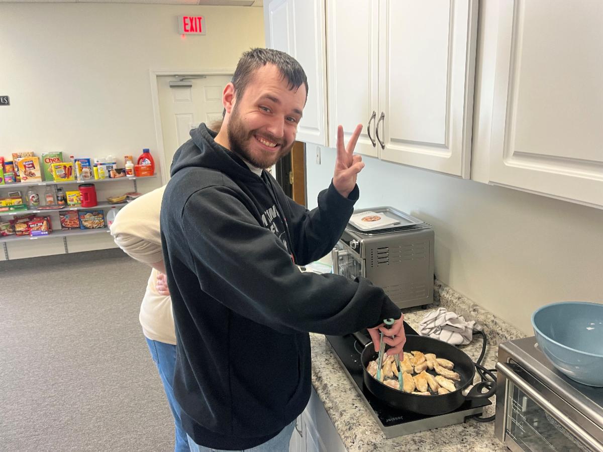 A young man smiles and makes the peace sign with his hand while sauteing chicken.