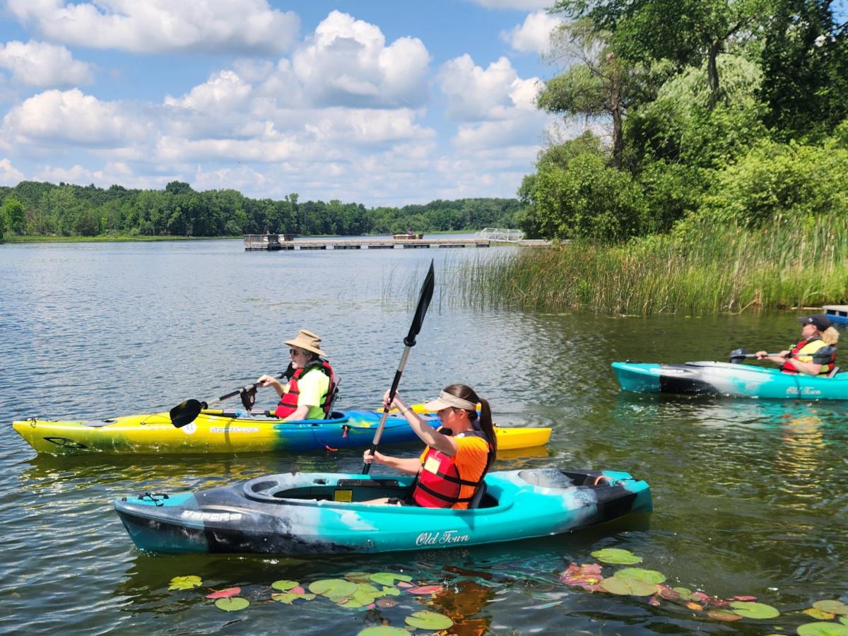 3 people wearing lifejackets are kayaking on a lake. One kayak has outriggers and a paddle pivot.