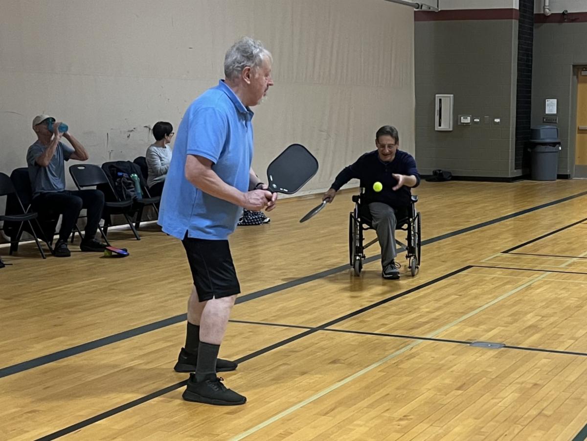 Two men play pickleball in a gym. One man is standing, ready to return a serve while another man in a wheelchair hits the ball with a paddle. Other players and observers sit along the wall in the background.