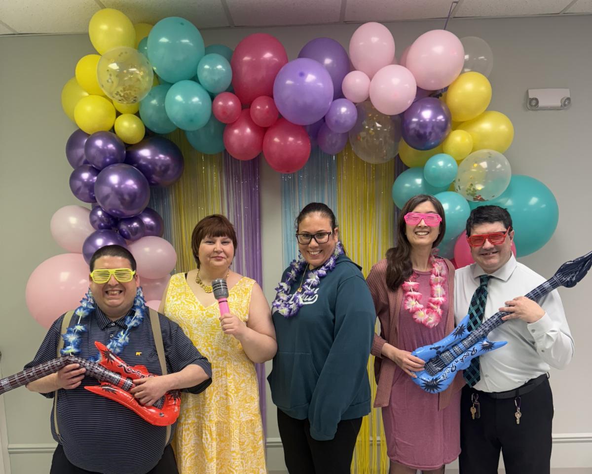 Five smiling people at a spring karaoke dance pose in front of a balloon arch and streamers, wearing sunglasses and leis and holding a inflatable microphone and guitars. 