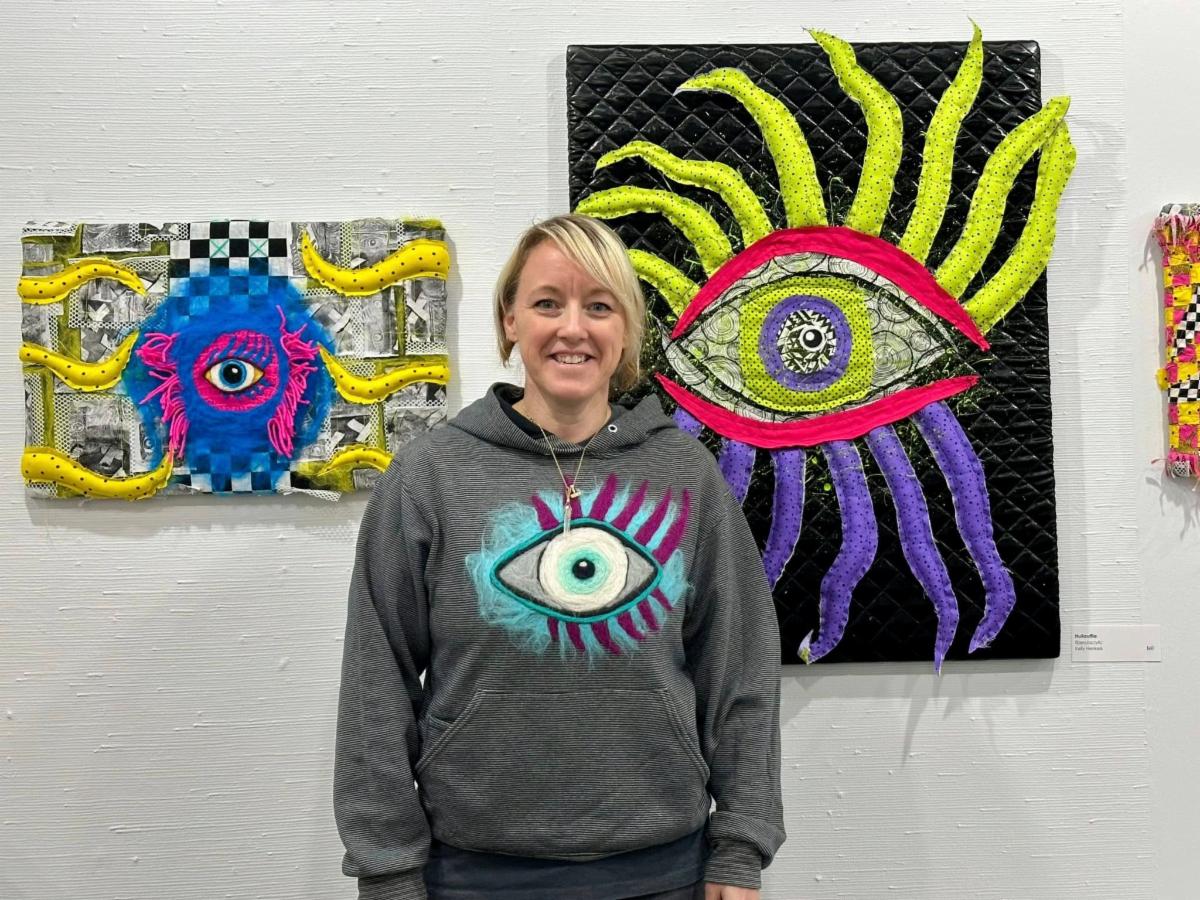 Artist Kelly Henkels standing in front of 2 fiber arts hall pieces and wearing a shirt which she needle felted an eye