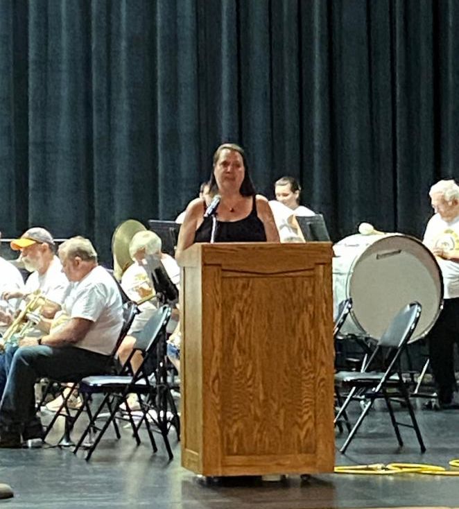SMAC Executive Director Nicole DeBoer stands at a podium with orchestra members behind her.