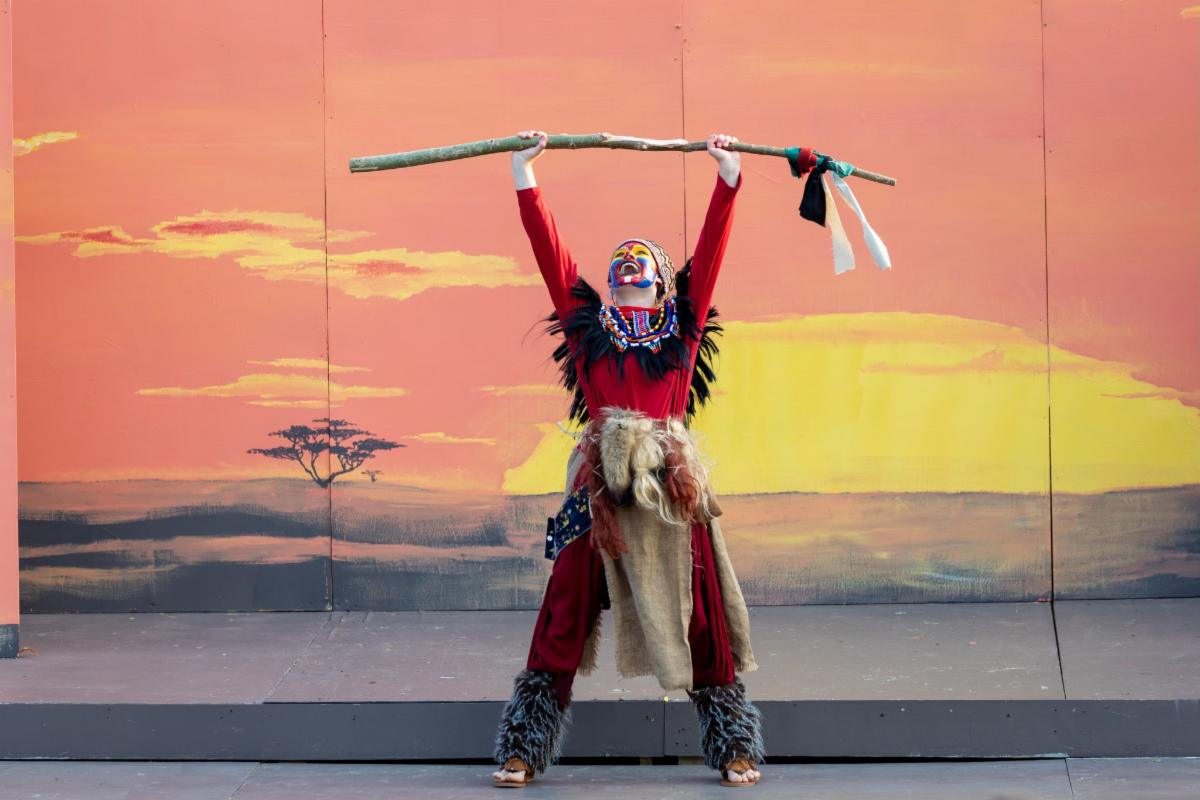 Youth performer in the Lion King Musical holds a staff high with a sunset painted background